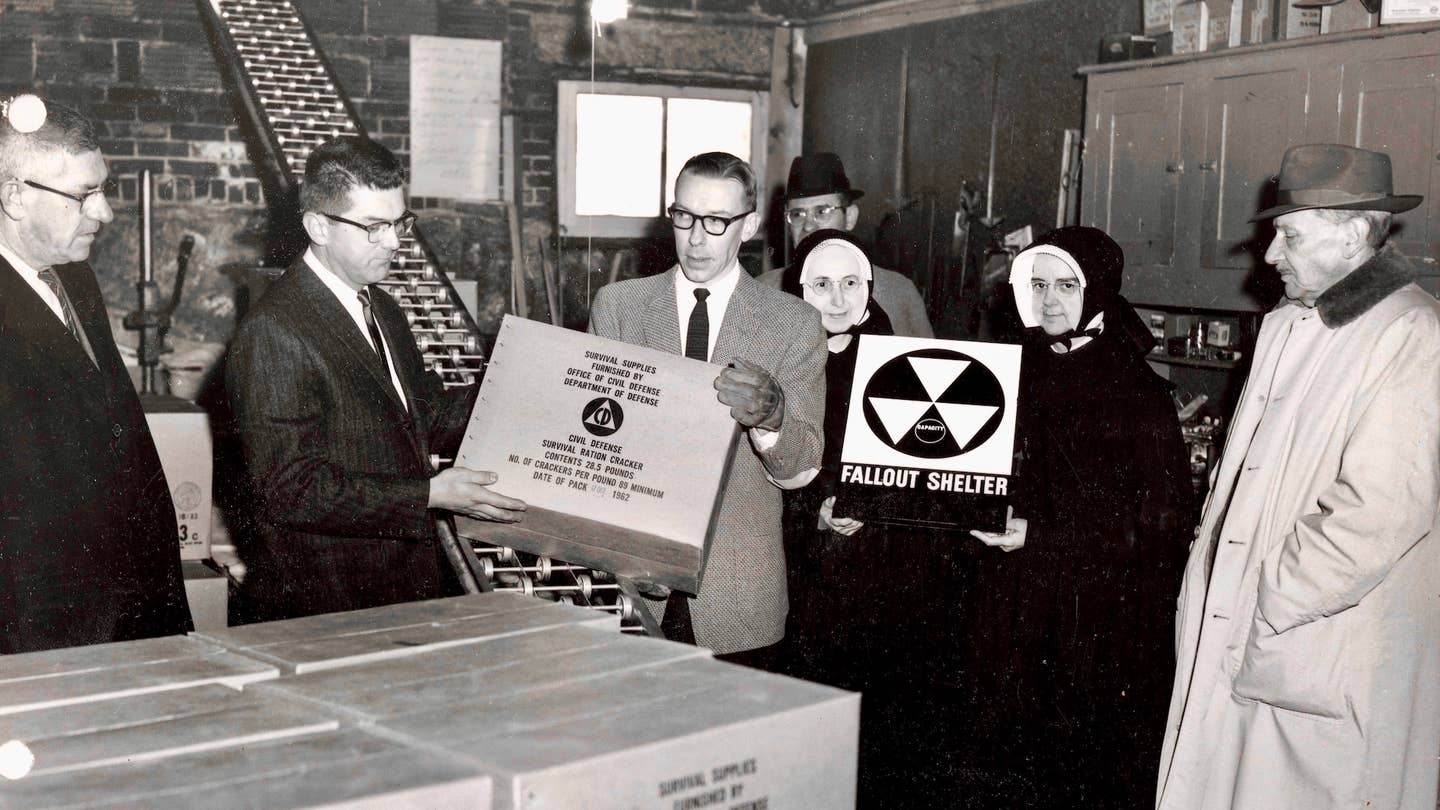 This photograph depicts a fallout shelter being furnished with Civil Defense survival rations at Villa Augustina Academy in Goffstown, New Hampshire. Mother Wilfred and Mother Superior Liguori of the Religious of Jesus and Mary, the religious order that founded and operated the school, appear in the photograph, holding a fallout shelter sign.